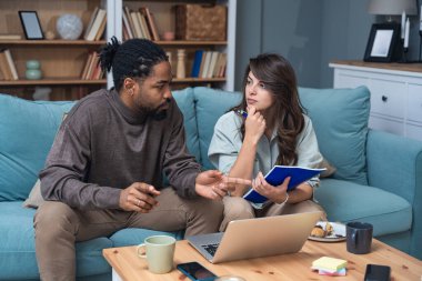 Genç çift evlerinde uzaktan çalışıyor, belge ve verileri analiz ediyor, rahat modern oturma odasında dizüstü bilgisayar kullanıyor, mali raporları ve iş stratejisini tartışıyor. Birkaç hedef, basit yaşam desteği..