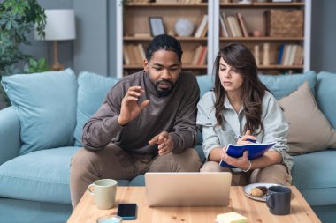 Genç çift evlerinde uzaktan çalışıyor, belge ve verileri analiz ediyor, rahat modern oturma odasında dizüstü bilgisayar kullanıyor, mali raporları ve iş stratejisini tartışıyor. Birkaç hedef, basit yaşam desteği..