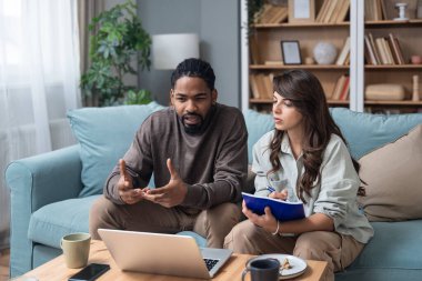 Genç çift evlerinde uzaktan çalışıyor, belge ve verileri analiz ediyor, rahat modern oturma odasında dizüstü bilgisayar kullanıyor, mali raporları ve iş stratejisini tartışıyor. Birkaç hedef, basit yaşam desteği..