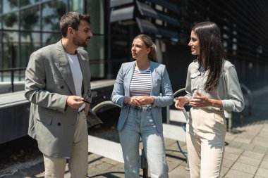 Kurumsal profesyonellerden oluşan çeşitli bir ekip dışarıda iş birliği yapıyor, modern cam ofis binası önünde dizüstü bilgisayardaki verileri analiz ederken stratejik tartışmalara girişiyorlar..