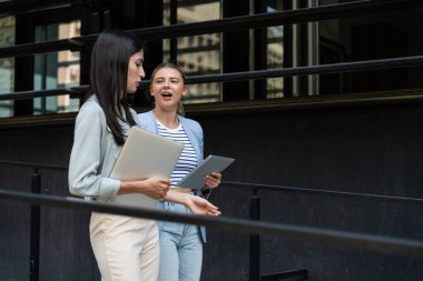 Kurumsal profesyonellerden oluşan çeşitli bir ekip dışarıda iş birliği yapıyor, modern cam ofis binası önünde dizüstü bilgisayardaki verileri analiz ederken stratejik tartışmalara girişiyorlar..