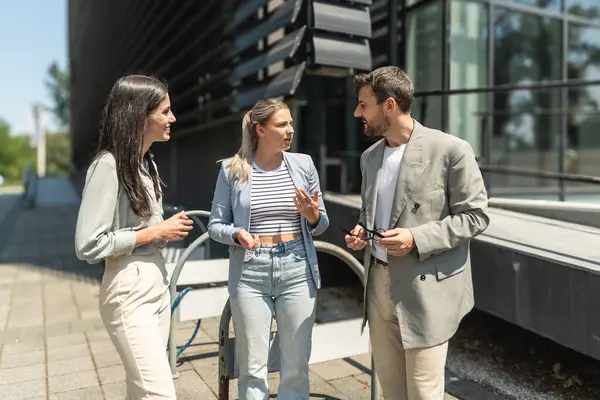 Kurumsal profesyonellerden oluşan çeşitli bir ekip dışarıda iş birliği yapıyor, modern cam ofis binası önünde dizüstü bilgisayardaki verileri analiz ederken stratejik tartışmalara girişiyorlar..