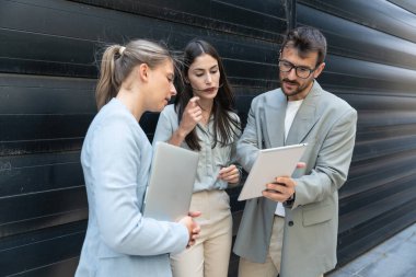 Modern cam ofis binası önünde dizüstü bilgisayardaki verileri analiz ederken dışarıda iş birliği yapan kurumsal profesyonellerden oluşan çeşitli iş adamları..