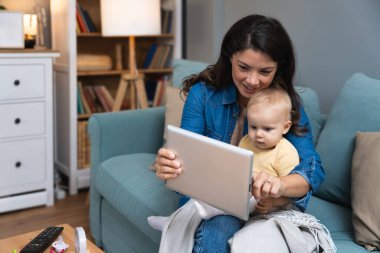 Gülümseyen genç anne evde tabletle meraklı bir kızı eğlendiriyor. Parlak oturma odasındaki rahat koltukta birlikte oturuyorlar. Ekran zamanı ve oynak dijital etkileşimler sayesinde birbirlerine bağlanıyorlar.