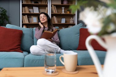 Mutlu genç bir kadın rahat bir kanepeye uzanıp ilginç bir kitap okuyor ve kulaklıkla müzik dinliyor, evde boş vakitlerin tadını çıkarıyor. Basit yaşam hedefleri, hafta sonu aktiviteleri konsepti.