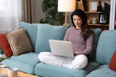 Konforlu oturma odasında dizüstü bilgisayar kullanan odaklanmış genç bir kadın. Serbest çalışan kadın, profesyonel iç mimar. İnterneti yeni fikirler ve müşteriler için ilham için düşünüyor ve kullanıyor..