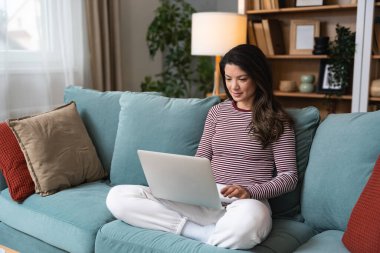 Konforlu oturma odasında dizüstü bilgisayar kullanan odaklanmış genç bir kadın. Serbest çalışan kadın, profesyonel iç mimar. İnterneti yeni fikirler ve müşteriler için ilham için düşünüyor ve kullanıyor..
