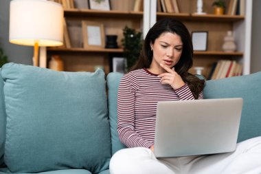 Konforlu oturma odasında dizüstü bilgisayar kullanan odaklanmış genç bir kadın. Serbest çalışan kadın, profesyonel iç mimar. İnterneti yeni fikirler ve müşteriler için ilham için düşünüyor ve kullanıyor..