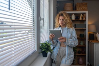 Rahat pijamalı genç bir kadın elinde dijital tablet olan parlak pencerenin yanında duruyor. Uzaktan çalışma, ders çalışma, mesajlar okuma, internette sohbet etme, flört etme, yalnızlığın tadını çıkarma..