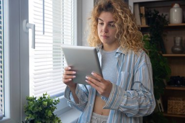 Rahat pijamalı genç bir kadın elinde dijital tablet olan parlak pencerenin yanında duruyor. Uzaktan çalışma, ders çalışma, mesajlar okuma, internette sohbet etme, flört etme, yalnızlığın tadını çıkarma..