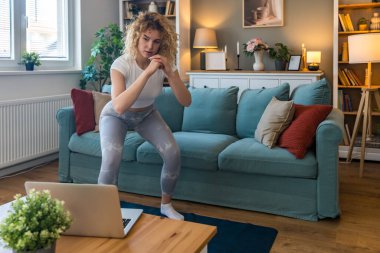 Spor kıyafetli genç bir kadın evde spor yapıyor, bilgisayar ekranından sanal antrenörü takip ediyor, çevrimiçi egzersiz oturumuna odaklanıyor, rutin, motivasyon ve sağlıklı yaşam tarzını sürdürüyor.