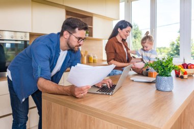 Neşeli genç aile, modern mutfak anne ve kızıyla kaynaşıyor. Tezgahta oynarken gülüyorlar. Babası uzak işlere odaklanıyor, ebeveynlik, sevgi ve teknolojiyi harmanlıyor. Basit yaşam.