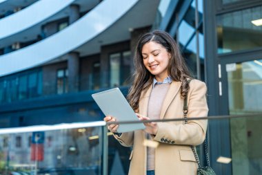 Modern ofis binasının dışında kendine güvenen profesyonel bir kadın dijital tabletle etkileşimde bulunuyor, dinamik bir iş ortamında odak, yenilik ve kentsel kurumsal yaşam tarzını gösteriyor.