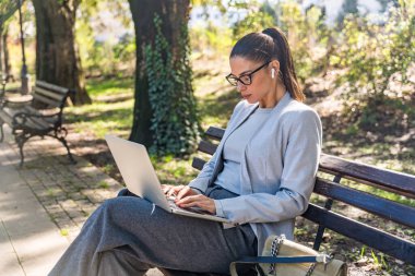 Parktaki bankta oturan profesyonel meslekten bir kadın, açık havada rahatlamayı verimlilikle harmanlamaya odaklanmış modern dijital göçebe yaşam tarzı ve kentsel sükuneti sergiliyor.