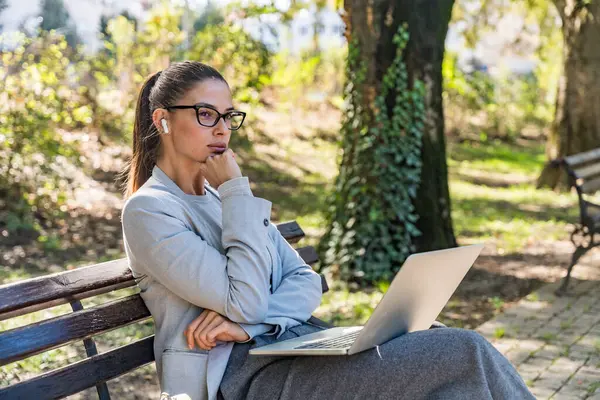 Parktaki bankta oturan profesyonel meslekten bir kadın, açık havada rahatlamayı verimlilikle harmanlamaya odaklanmış modern dijital göçebe yaşam tarzı ve kentsel sükuneti sergiliyor.