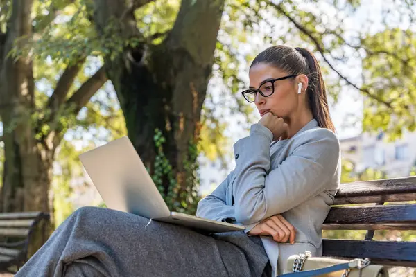Parktaki bankta oturan profesyonel meslekten bir kadın, açık havada rahatlamayı verimlilikle harmanlamaya odaklanmış modern dijital göçebe yaşam tarzı ve kentsel sükuneti sergiliyor.