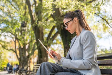 Parkta oturan genç iş kadını, dijital tablet üzerinde çalışıyor, profesyonel meslekte çalışıyor, ofis dışında kalabalık toplanıyor ve işine odaklanıyor. Finans ve ekonomi uzmanı tele-çalışma