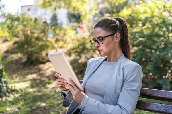 Parkta oturan genç iş kadını, dijital tablet üzerinde çalışıyor, profesyonel meslekte çalışıyor, ofis dışında kalabalık toplanıyor ve işine odaklanıyor. Finans ve ekonomi uzmanı tele-çalışma