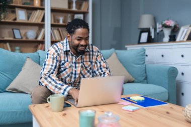 Genç girişimci evden çalışıyor, bilgisayarında önerilerini okurken plan yapıyor, rahat bir oturma odasında, modern yaşam tarzında ve uzak iş konseptinde başarılı bir anın tadını çıkarıyor..