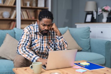 Genç girişimci evden çalışıyor, bilgisayarında önerilerini okurken plan yapıyor, rahat bir oturma odasında, modern yaşam tarzında ve uzak iş konseptinde başarılı bir anın tadını çıkarıyor..