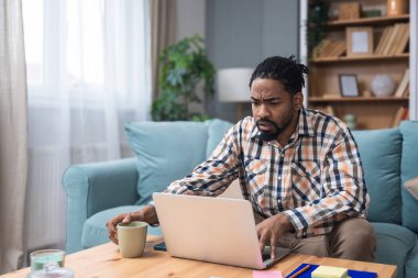 Genç girişimci evden çalışıyor, bilgisayarında önerilerini okurken plan yapıyor, rahat bir oturma odasında, modern yaşam tarzında ve uzak iş konseptinde başarılı bir anın tadını çıkarıyor..