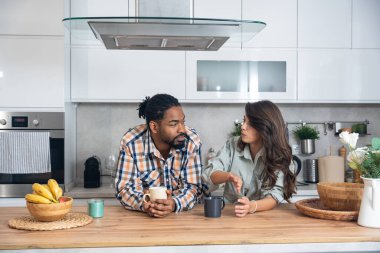 Mutlu çift yeni evlerinin mutfağında kahvenin tadını çıkarıyorlar. Birlikte rahat bir sabah geçiriyorlar. Rahatlamış ve gülümsüyorlar.