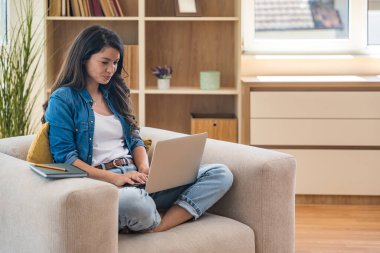 Evden uzakta çalışan genç bir iş kadını, rahat bir koltukta dizüstü bilgisayarla oturuyor. Modern çalışma alanı, dijital iş, rahat ortam, ve esnek online kariyer fırsatı