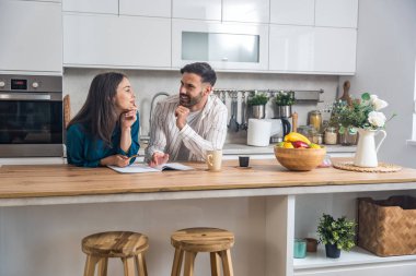 Genç nişanlı çift modern mutfaklarında bir arada duruyorlar, düğün için konuk listesi hazırlıyorlar, yemek menüleri düzenliyorlar, odaklanıyorlar ve gülümsüyorlar.