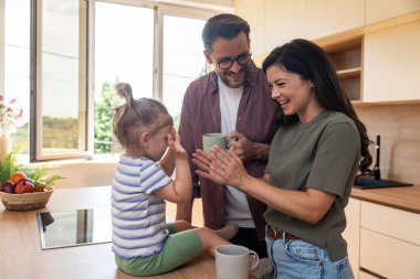 Mutlu ebeveynler mutfakta birlikte huzurlu bir sabah geçirirken neşeli küçük kızları tezgahta gülümseyip oyuncaklarla oynarken kahve içiyorlar. Sakin, sıcak aile anları..