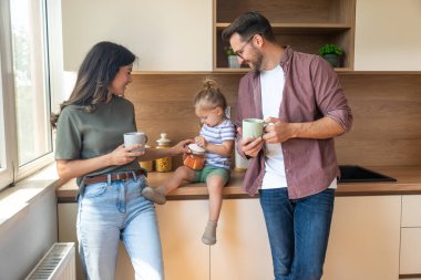 Mutlu ebeveynler mutfakta birlikte huzurlu bir sabah geçirirken neşeli küçük kızları tezgahta gülümseyip oyuncaklarla oynarken kahve içiyorlar. Sakin, sıcak aile anları..