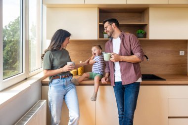 Mutlu ebeveynler mutfakta birlikte huzurlu bir sabah geçirirken neşeli küçük kızları tezgahta gülümseyip oyuncaklarla oynarken kahve içiyorlar. Sakin, sıcak aile anları..