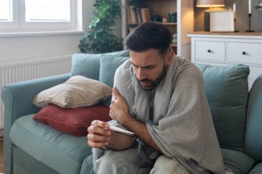 Battaniyeye sarılı yatan ve evde sıcaklığı ölçen soğuk algınlığı sırasında yüksek ateş, zayıflık ve fiziksel rahatsızlık hisseden ve grip belirtilerini atlatmaya çalışan hasta bir adam.