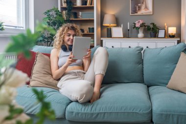 Genç bir kadın ya da kız elinde dijital tabletle kanepede oturmuş, kablosuz kulaklıkla çalacak müzik arıyor. Keyifli, mutlu bir kadın eğlence ya da online flört için yayın servisi seçiyor..