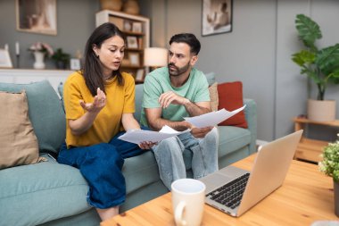 Evdeki kanepede oturan endişeli genç çift, dizüstü bilgisayar ve kağıttaki mali belgeleri ve istatistikleri analiz ederken, beklenmedik harcamalar, borç veya olumsuz bütçe raporu sonuçlarından endişe duyuyor.