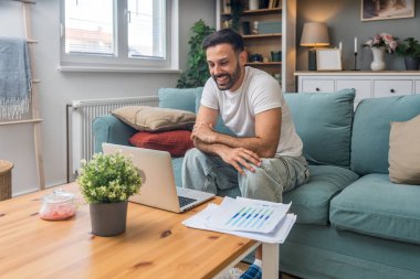 Genç iş adamı küçük bir şirket sahibi, evde çalışıyor, grafikleri ve dizüstü bilgisayarları kontrol ediyor. Yeni ürün stratejisinin maliyetlerini ve giderlerini hesaplayan yenilikçi genç serbest erkek.