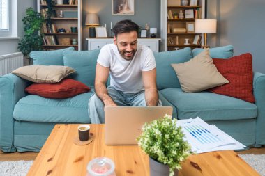 Genç iş adamı küçük bir şirket sahibi, evde çalışıyor, grafikleri ve dizüstü bilgisayarları kontrol ediyor. Yeni ürün stratejisinin maliyetlerini ve giderlerini hesaplayan yenilikçi genç serbest erkek.