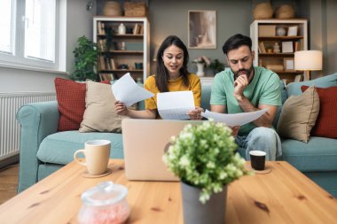 Evdeki kanepede oturan endişeli genç çift, dizüstü bilgisayar ve kağıttaki mali belgeleri ve istatistikleri analiz ederken, beklenmedik harcamalar, borç veya olumsuz bütçe raporu sonuçlarından endişe duyuyor.