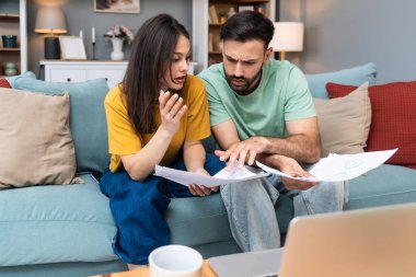 Evdeki kanepede oturan endişeli genç çift, dizüstü bilgisayar ve kağıttaki mali belgeleri ve istatistikleri analiz ederken, beklenmedik harcamalar, borç veya olumsuz bütçe raporu sonuçlarından endişe duyuyor.
