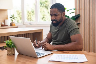 Milenyum Afrikalı Amerikalı adam Webinar ya da iş toplantısı sırasında not alıyor, dizüstü bilgisayar kullanıyor, çalışıyor ya da internetten çalışıyor, ofis masasında oturuyor. Serbest iş, e-ticaret girişimi.