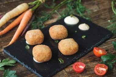 delicious fried mozzarella cheese balls served on a black ceramic plate, fresh vegetables on a wooden rustic table