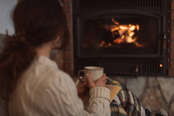Young woman sitting at home by the fireplace with a mug of hot coffee or tea and a book, she is wearing woollen sweater. Cold houses with low temperatures in Europe concept. Selective focus on a mug. 