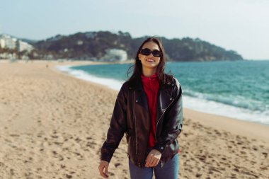 Plajda duran siyah güneş gözlüklü ve deri ceketli şık bir modelin portresi. Genç, seksi, tatilde gülümseyen kadın deniz esintisinin ve kameraya bakmanın tadını çıkarıyor. Çekici, güzel kız..