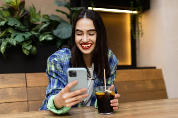 Happy zoomer multiethnic teen girl checking social media holding smartphone at cafe. Smiling young  woman using mobile phone app playing game, shopping online, ordering delivery. Amazing toothy smile.
