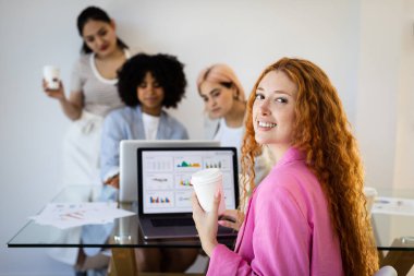 Genç, mutlu, kızıl saçlı bir kız elinde bir fincan kahveyle bir ofisin önünde oturuyor. Önünde, o çalışırken çizelgeleri ve grafikleri gösteren bir bilgisayar ekranı var..