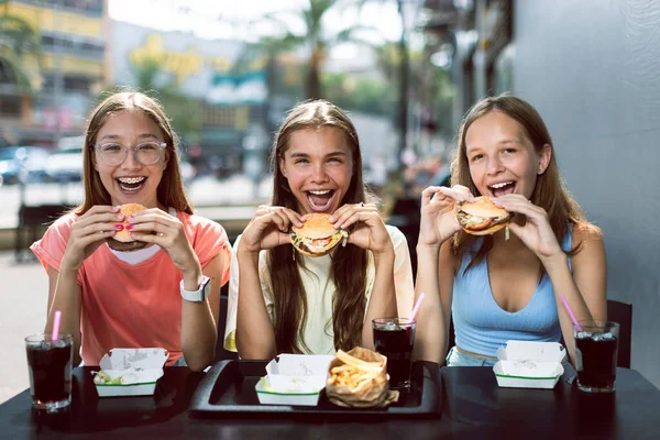 三个快乐的笑着的少女 15岁 坐在外面的咖啡馆里 享用汉堡包和可乐 新鲜饮料 年轻的女士拿着汉堡包 准备愉快地吃 — 图库照片