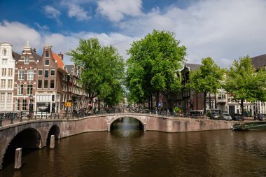 Amsterdam, Hollanda - 21 Mayıs 2025: Tuğla kemer köprüsü, ağaçlı bankalar ve mavi gökyüzü altındaki tarihi binaların yer aldığı Amsterdam kanal sahnesi
