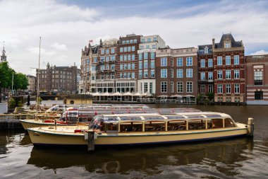 Amsterdam, Hollanda - 21 Mayıs 2025: Arka planda tarihi binalar ve Amsterdam kanalı boyunca demirlemiş çok sayıda cam çatılı kanal turu tekneleri
