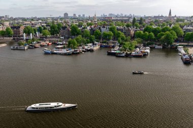 Amsterdam, Hollanda - 21 Mayıs 2025: Amsterdam 'ın arka planında Amsterdam şehri bulunan Amstel Nehri' nin hava panoramik manzarası, nehir botlarla benekli ve ağaçlarla çevrili ve uzak şehir buğdaylarıyla çevrili