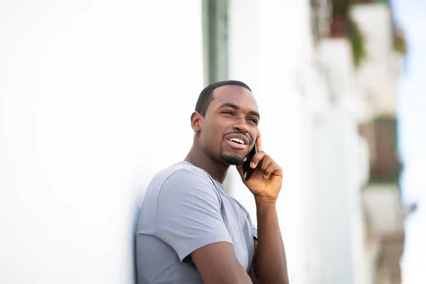 携帯電話で話しているハンサムな若いアフリカ系アメリカ人の男の肖像画は外の白い壁にもたれて — ストック写真