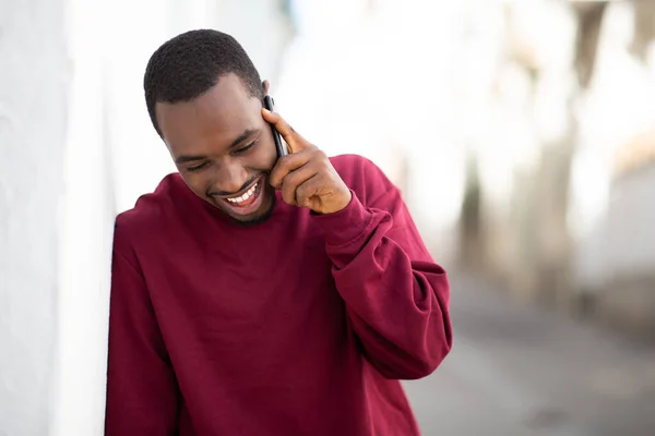Portrait of smiling young black man leaning to wall and talking on mobile phone outside